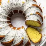 Cake au citron et pavot fraîchement cuit, décoré de zestes de citron et d’un léger glaçage, posé sur une table en bois avec des tranches de citron et des graines de pavot.